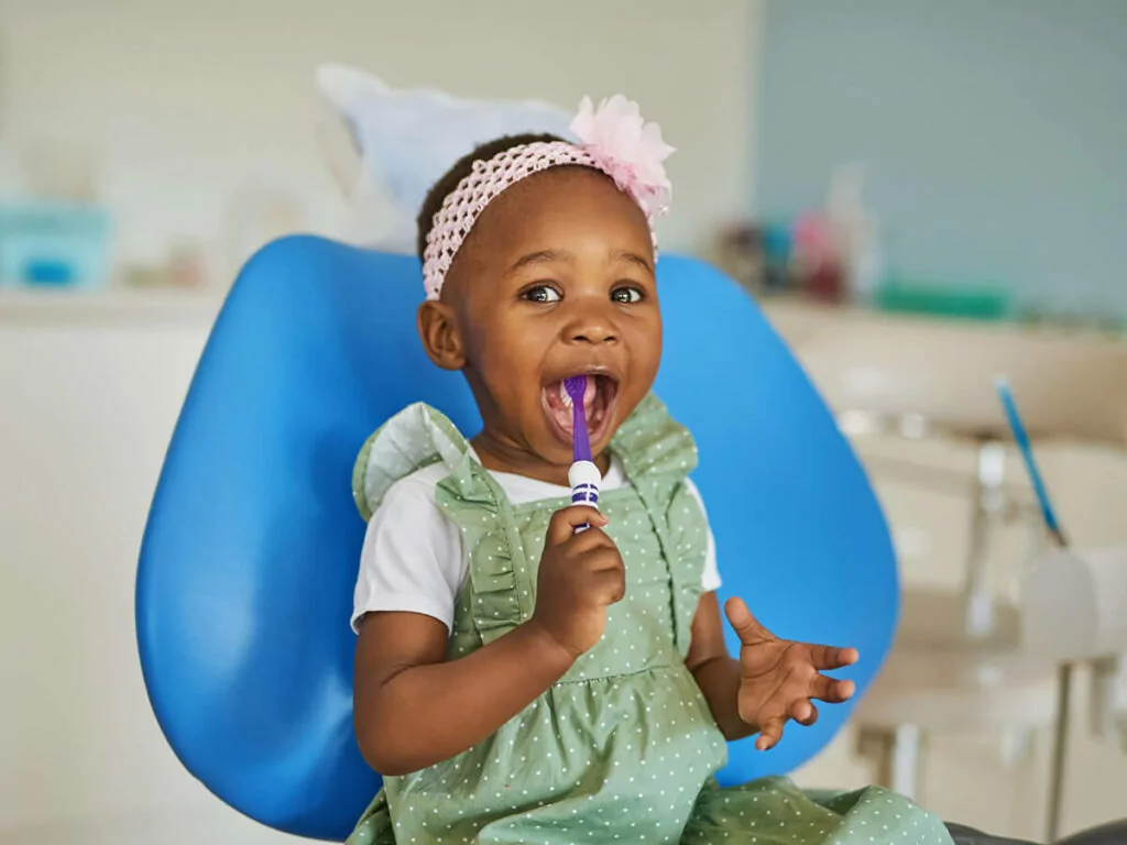 The image shows a young child sitting on a blue chair, in a dental office. The child is wearing a green polka-dotted dress with a white shirt underneath and a pink headband with a flower. The child is holding a toothbrush.