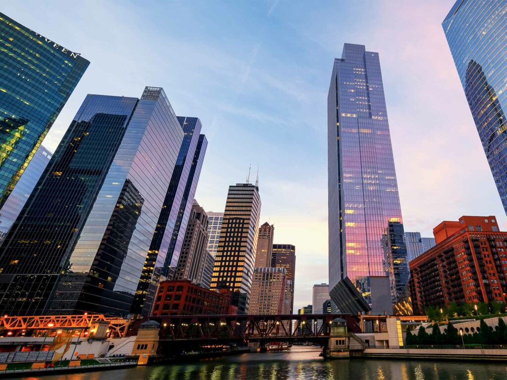 Cityscape with tall skyscrapers reflecting light, river with a bridge in the foreground.