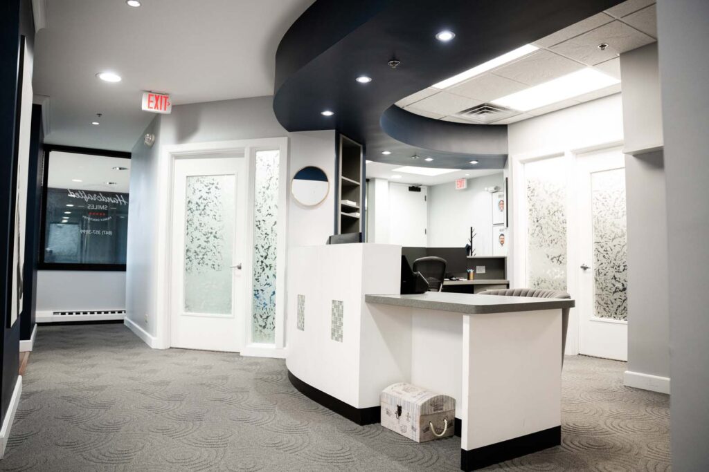 Interior view of Handcrafted Smiles dental office in Arlington Heights, IL, featuring a sleek front desk with a modern design. The office has a curved, dark ceiling detail with embedded lights, decorative frosted glass doors, and a patterned carpet. A small seating area with contemporary decor is visible in the background.