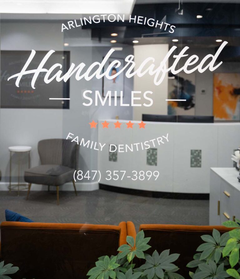 Glass entrance of Handcrafted Smiles dental office, prominently displaying the office name 'Handcrafted Smiles - Family Dentistry' in elegant white script on the door. The view through the glass shows a modern reception area with stylish seating and the office phone number.