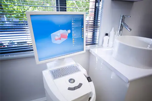 A dental CAD/CAM (Computer-Aided Design and Computer-Aided Manufacturing) system in a dental office. The setup includes a computer with a monitor displaying a 3D model of a dental restoration, and a milling machine used to fabricate the dental restoration based on the digital design.