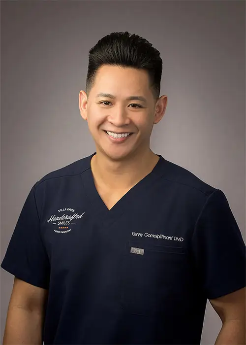 Close up image of Dr. Kenny Gomolplitinant wearing a navy blue scrub while his one hand at a pocket. The background is mix of dark and light color.